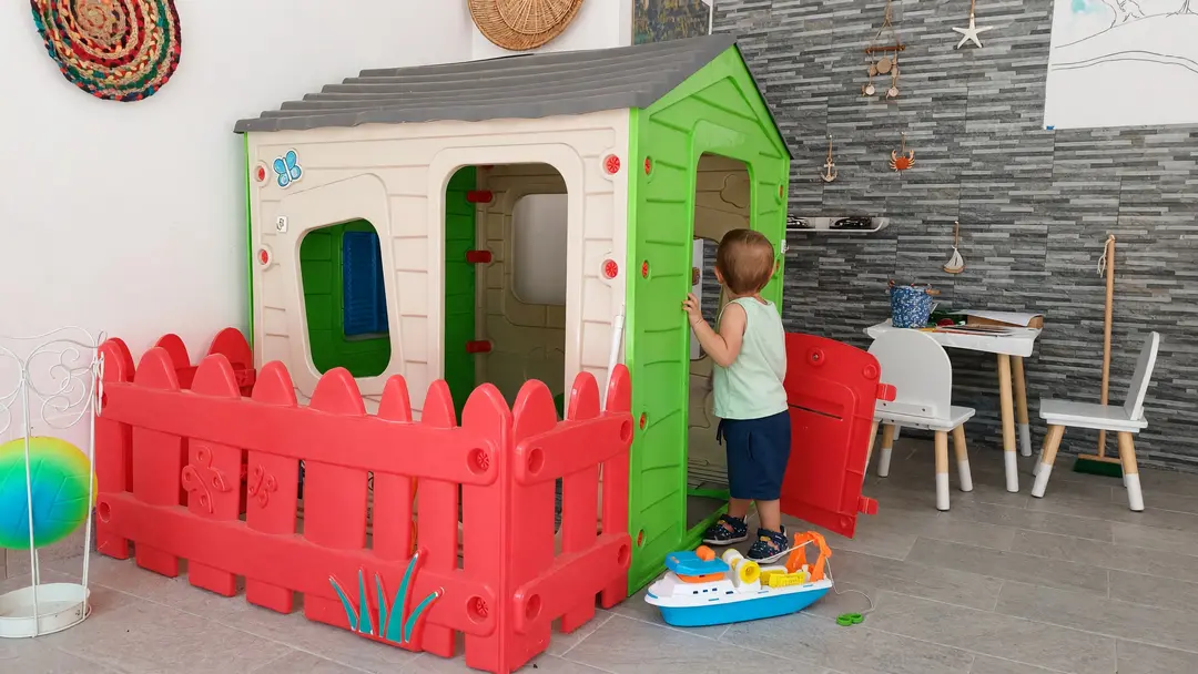 Un bimbo alla porta della casetta dei bambini, sotto il portico, con lo steccato rosso e una barca giocattolo