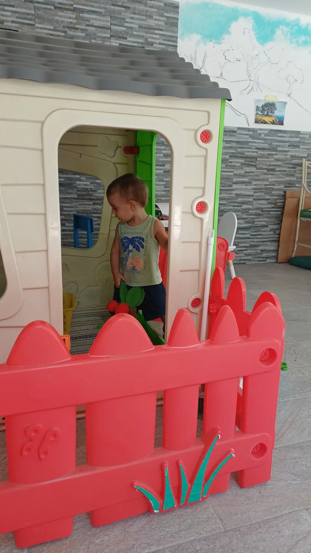 Un bimbo gioca dentro la casetta dei bambini, visto dalla porta, oltre lo steccato rosso