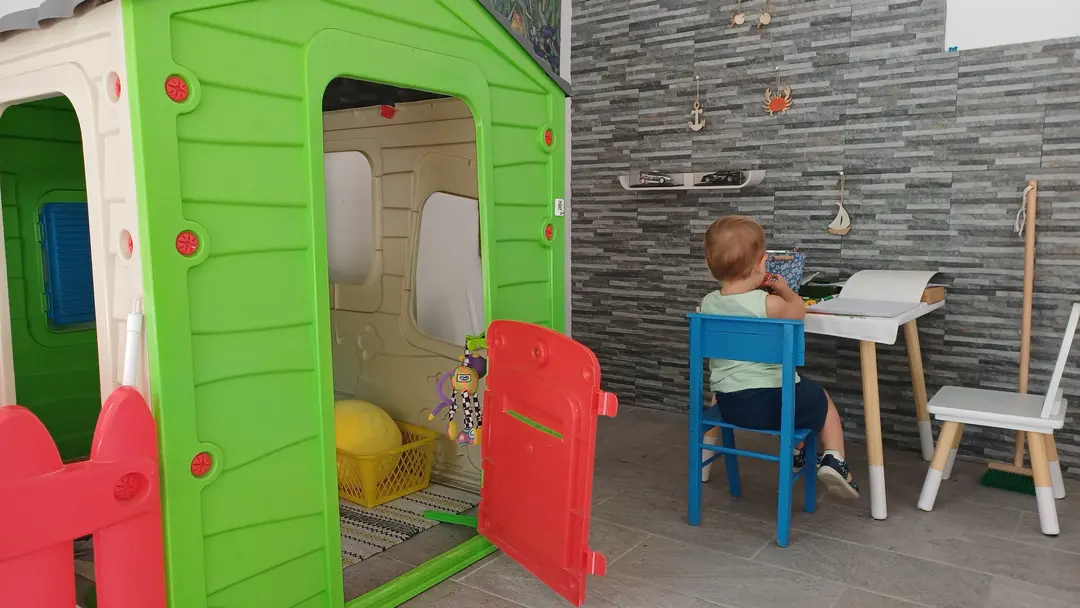 Un bimbo di spalle, seduto sulla seggiolina blu al tavolino da disegno, accanto alla casetta dei bambini con la porta aperta